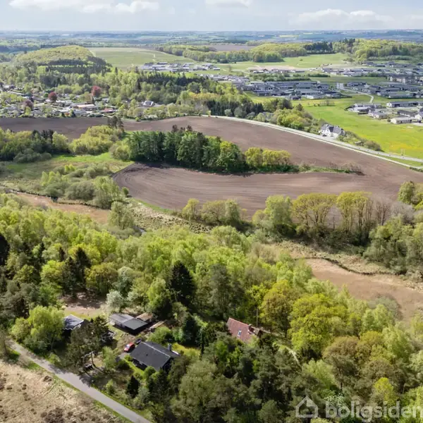 Luftfoto viser landbrug og skove; marker strækker sig mellem tætte skovområder. I baggrunden ses en by med veje og beboelsesområder.