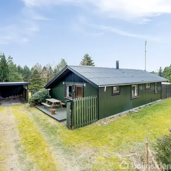 Et grønt træhus står på en græsklædt grund omgivet af træer. Der er en terrasse med en bænk foran huset og en redskabsbygning til venstre i billedet.