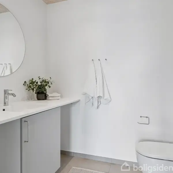 Rundt spejl hænger over en moderne vask med toiletsager og plante, mens et toilet er placeret ved siden af i det hvide badeværelse med håndklæder på væggen.