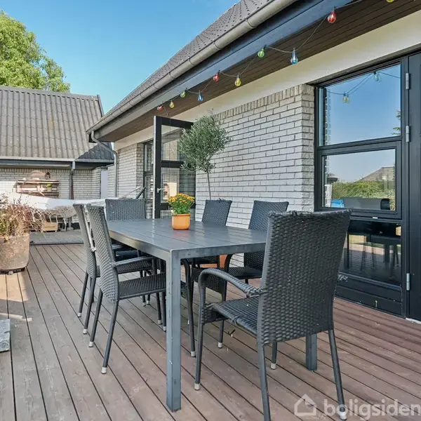 Terrassestolene og bordet står stationære på en træterrasse foran et hvidt murstenshus. I baggrunden ses en grill og farverige lyskæder. Græsplæne og planter omgiver terrassen.