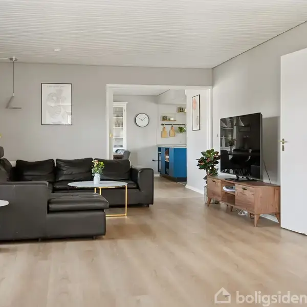 Sort lædersofa står i lys stue med trægulv, ved siden af sofaen er der et lille rundt bord. Foran står et tv på et træbord. Dør til køkken er åben.