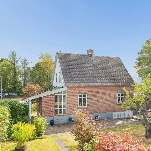 Murstenshus med spidst tag står stille i et grønt haveområde. Træer og buske omgiver huset, og en sti fører til hoveddøren. Sollys skinner på haven.