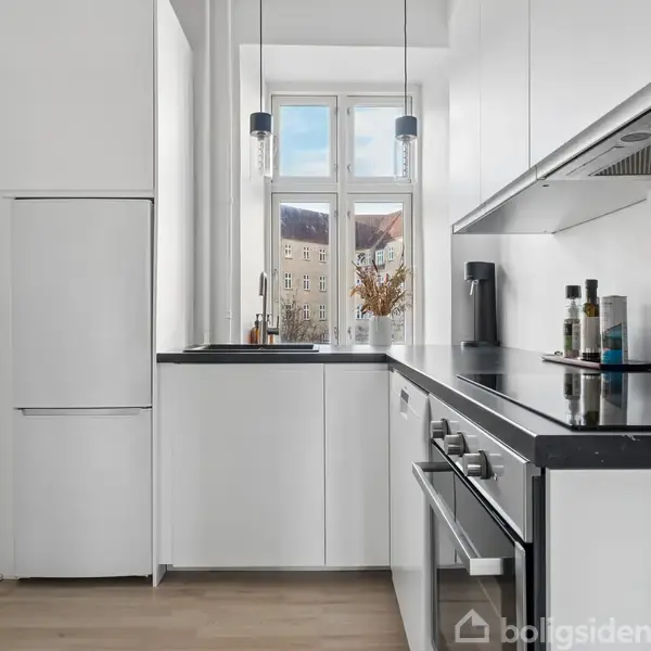 Et hvidt, moderne køkken med indbyggede skabe og apparater. Der er en ovn i sort og stål, samt en hvid køleskab i venstre hjørne. Lyset strømmer ind gennem et vindue i baggrunden.