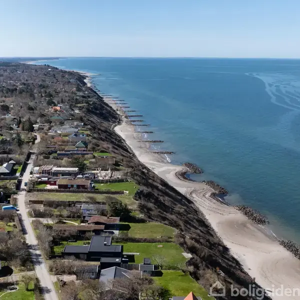 Kystlinje strækker sig parallel med en række huse og grønne områder. Havet ligger til højre, mens husene ligger til venstre langs en vej. Himlen er klar og blå.
