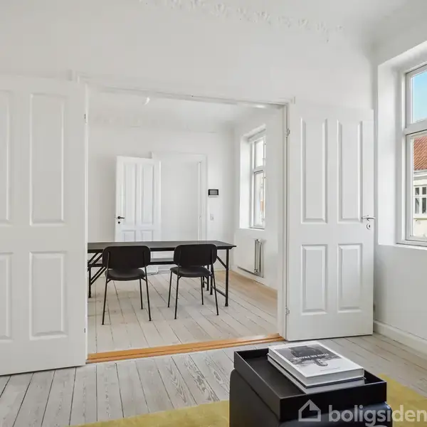 Et lyst rum med hvide vægge og gulve. Dobbelt døre åbner til et andet værelse med et sort bord og to stole. Store vinduer slipper naturligt lys ind.