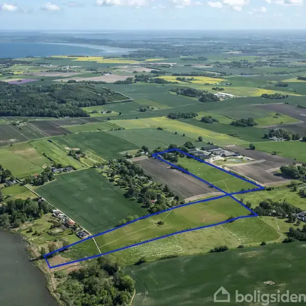 Landområde markeret med blå linje omfatter marker og en skovklædt kystlinje. Området ligger i et landbrugsområde med spredte huse og en sø i nærheden.