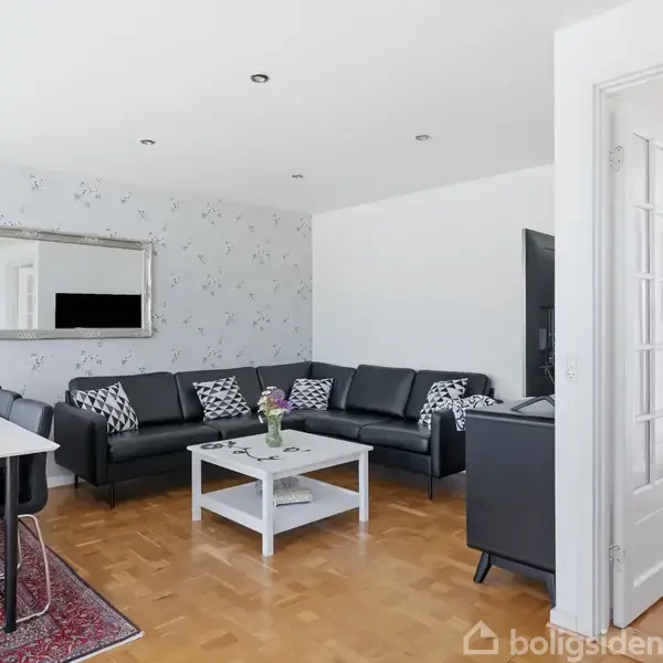 En sort hjørnesofa står i en stue med lyst trægulv og blomsterpræget tapet. En hvid sofabord og spisebord med stole står nær et stort spejl.