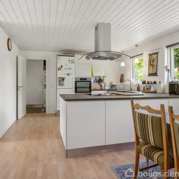 Køkkenø i lys, moderne stil med indbygget komfur og emhætte; omgivet af spisebord og stole. Store vinduer giver udsigt til grøn have. Trægulv og hvid loft beklædning.