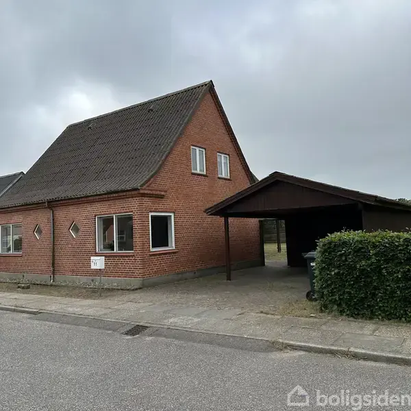Rødt murstenshus med skråt tag står stille ved vejen; til højre er en åben garage med en hæk ved siden af, under en grå skyfuld himmel.