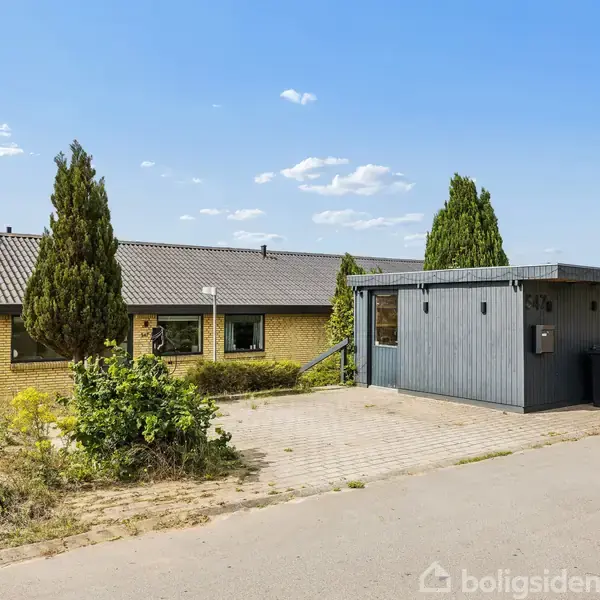 Et gult murstenshus med en gråmalet tilbygning står stille i et velplejet område med græs og buske. Huset har en flisebelagt indkørsel og affaldscontainere ved siden af.