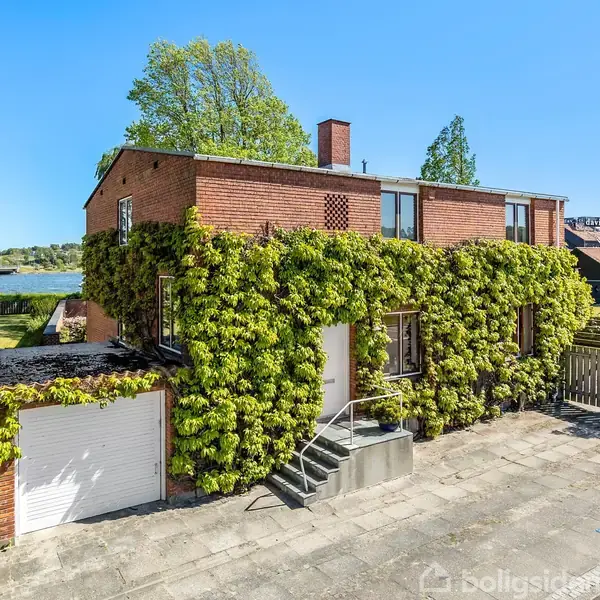 En murstensbygning dækket med klatreplanter står stille ved en søbred. Garagen er synlig til venstre, mens terrassen er omgivet af grønne træer og blå himmel.