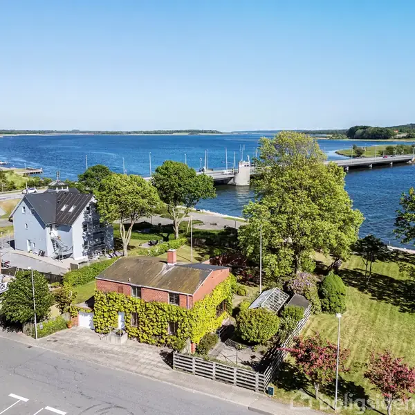 Et rødt hus med grønne klatreplanter omgiver facaden i en forstadshave. Det står tæt på en vej ved en sø med en bro og frodige træer i nærområdet.