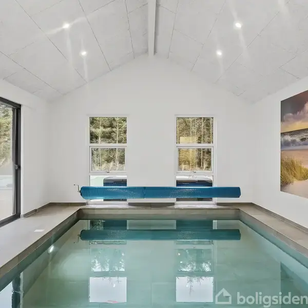 Indendørs swimmingpool med klart vand, hvor en blå rullegardin er monteret over. To vinduer bagved viser skovtræer udenfor. På højre væg hænger et stort billede af en solnedgang over en strand.