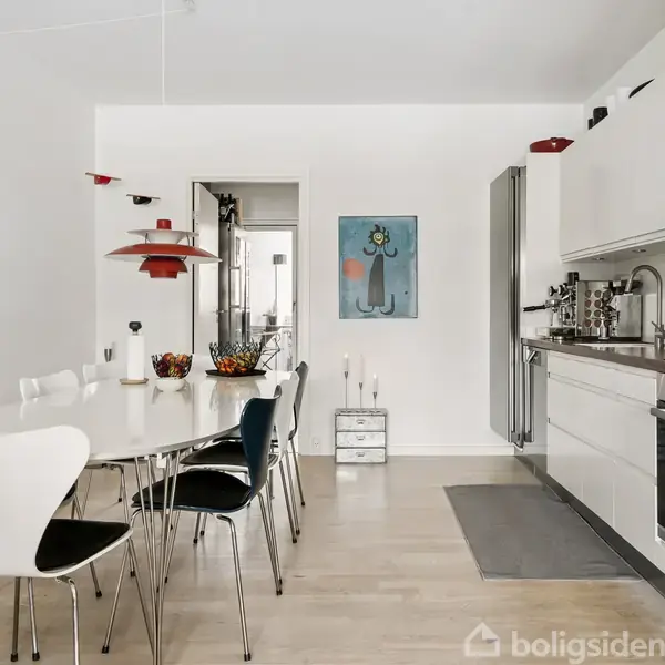 Et moderne køkken med hvid spisebord og stole midt i rummet. En rød lampe hænger over bordet. I baggrunden er et maleri på væggen og stilfuldt køkkenudstyr.