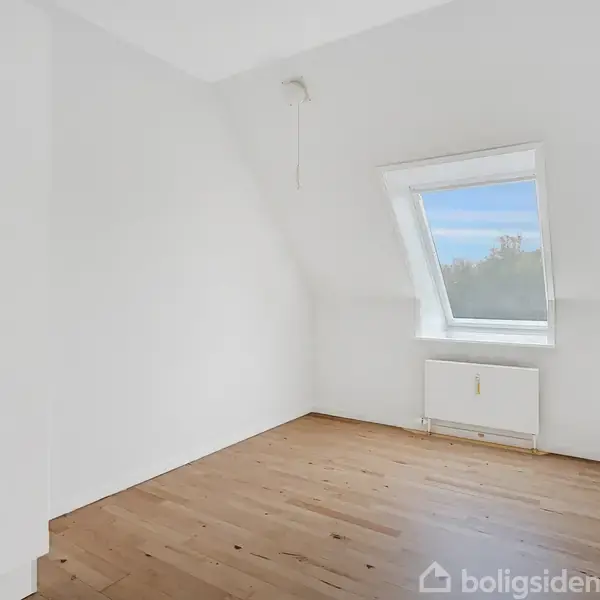 Et tomt rum med trægulv og hvidmalede vægge. Et stort skråtstillet vindue lader dagslys strømme ind, og der er en radiator placeret under vinduet. En hvid garderobe står til venstre.