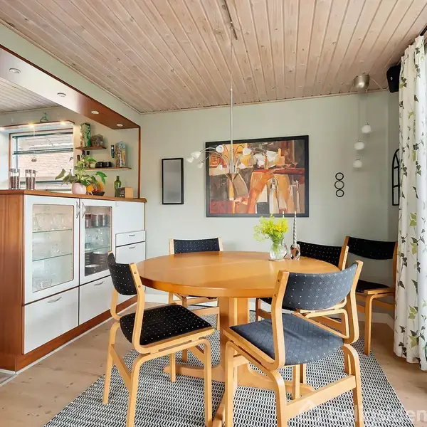 Et rundt spisebord med fem stole står i en spisestue. Der er en vase med blomster på bordet. Køkkenet ses i baggrunden, og en udsmykket væg med kunst hænger over bordet.