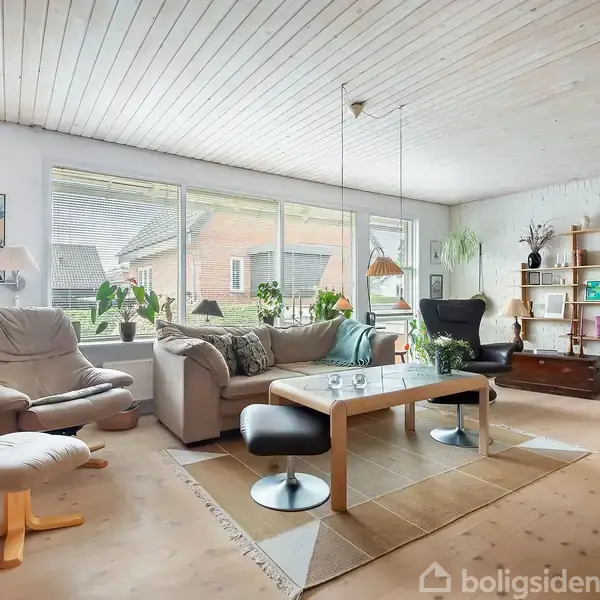Stue med sofagruppe og lænestole omkring et sofabord. Store vinduer lader lys ind, mens reoler og planter dekorerer rummet. Træloft og loftslys tilføjer en hyggelig atmosfære.