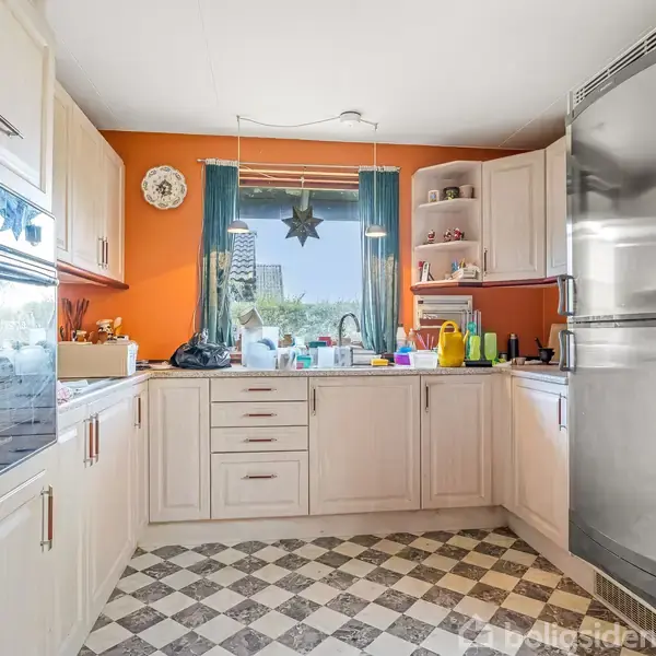 Et lyst køkken med hvide skabe og skuffer. Midt i rummet står en stålkomfur og et køleskab. Et vindue med grønne gardiner giver udsigt til en have.