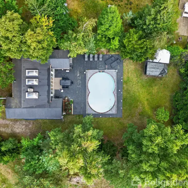 Hus med fladt tag og fem ovenlysvinduer omgivet af skov. Ved siden af ligger en hjerteformet pool på en terrasse. Området har tæt grøn beplantning.