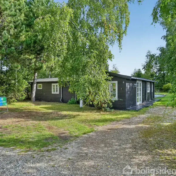 Et enkelt, mørkt træhus står omgivet af frodig bevoksning og grusvej foran. Et skilt ses nær indkørslen, der fører til huset.