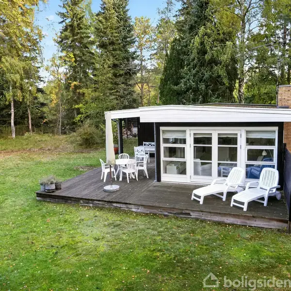 Huset er et moderne træhus med store vinduer og en terrasse. To liggestole og et bord med stole står udenfor. Huset er omgivet af frodig skov og grønne græsarealer.