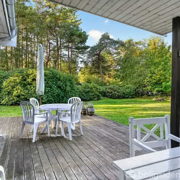 Træterrasse med hvidt havesæt og parasol står foran grønt græsareal og skov. En grå sky er placeret i baggrunden under en lyseblå himmel med enkelte skyer.