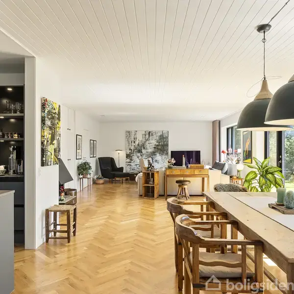 Spisebord med træstole og planter på toppen, placeret i et moderne spisekøkken. Store pendellamper hænger over bordet. I baggrunden ses en stue med sofagruppe og vinduer med udsigt.