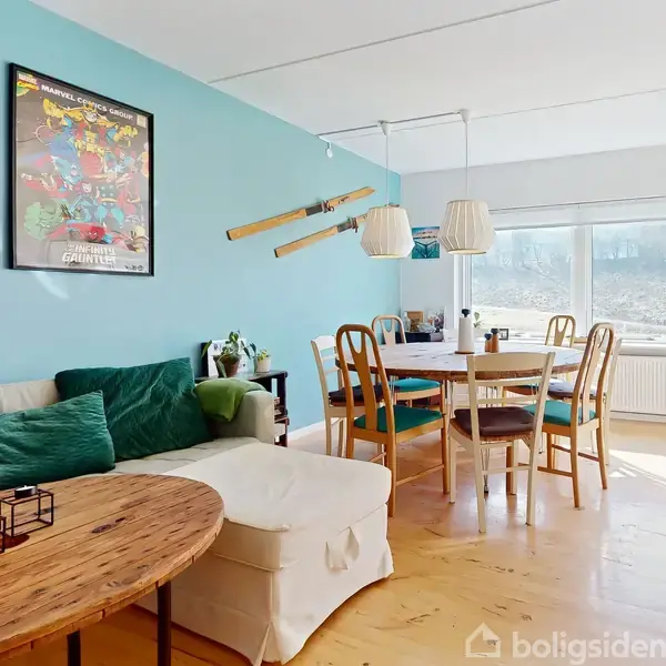 Sofa med grønne puder står ved et rundt træbord. En blå væg med et maleri og ski dekorerer stuen. Et spisebord med stole er tæt ved et stort vindue.
