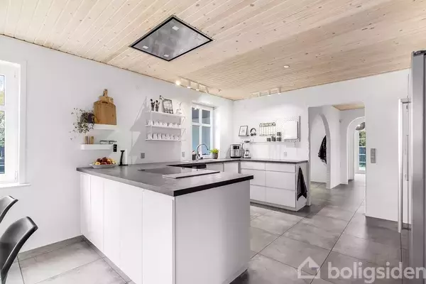 Køkkenø i moderne køkken, omgivet af hvide skabe og hylder. Der er en indbygget vask og frugtskål. Lyse trægulve og vindue med naturligt lysindfald.