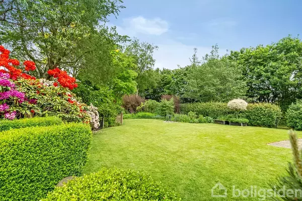 En velplejet have prydes af grønne buske og farverige blomster til venstre. Træer omkranser plænen, skaber en rolig atmosfære under en klar blå himmel.