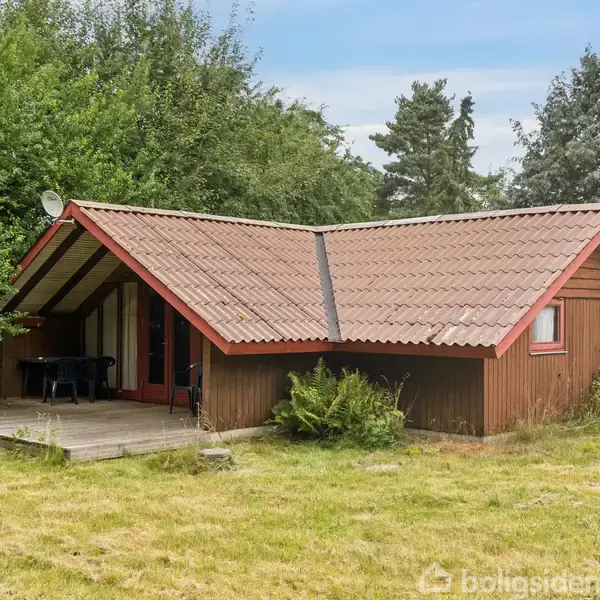 Træhus med skråtag står stille i en grøn have. En lille terrasse med bord og stole er til venstre, omgivet af buske og træer.