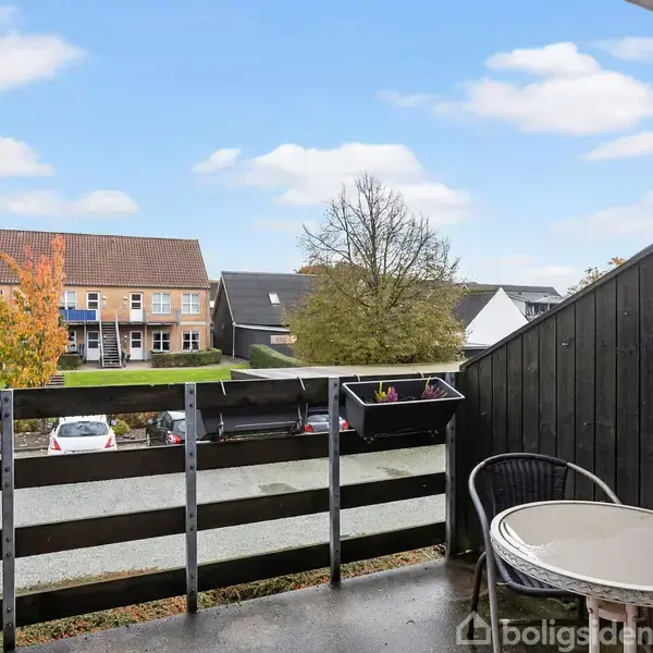 En altan med et rundt bord og en stol. Håndlisterne har blomsterkasser. Baggrunden viser et beboelseskvarter med rækkehuse, træer og parkerede biler under en blå himmel.