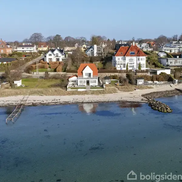 Hus med rødt tag står ved kysten, omgivet af en sandstrand og klart vand. Baggrunden har flere huse og grønne områder under en klar blå himmel.