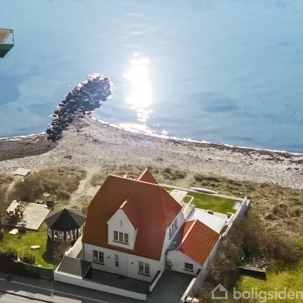 Hus med rødt tag ligger ved stranden. Sollys reflekteres i vandet. En stenmole og en træbro strækker sig ud i det klare, blå hav.