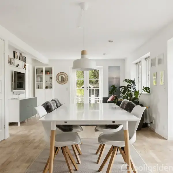 Et rummeligt spisebord med seks stole står på et lyst trægulv. Rummet har hvide vægge, moderne møbler og store vinduer, der lukker naturligt lys ind. Malerier pryder væggene.