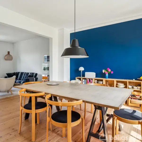 Spisebord med træstole står i et rum med trægulv, en blå væg og en hylde fyldt med tallerkener. En stue med sofaer ses i baggrunden.
