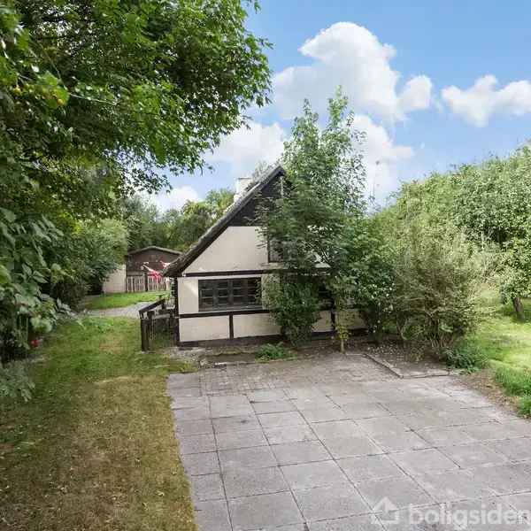 Et bindingsværkshus står omgivet af tæt bevoksning og græsplæner. Et flisebelagt område ses foran huset. Himlen er klar med spredte skyer.