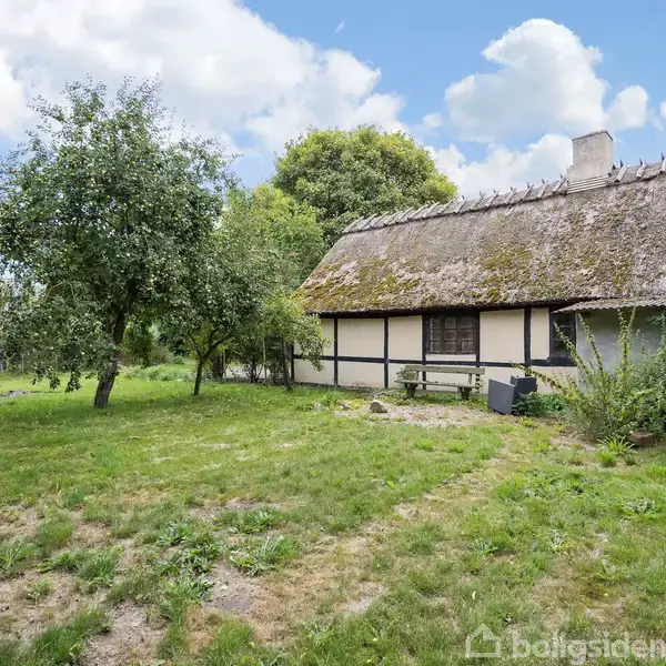 Et bindingsværkshus med stråtag står stille i en frodig have. Træerne omkring giver skygge, og himlen er delvist skyet, hvilket skaber et fredeligt landligt miljø.