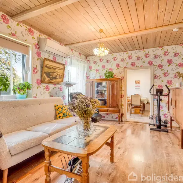 Sofa står i stue med blomstrede tapeter, foran et træbord med en vase på. Værelset har træloft, lysekrone og er dekoreret med planter og træmøbler, der skaber en hyggelig atmosfære.