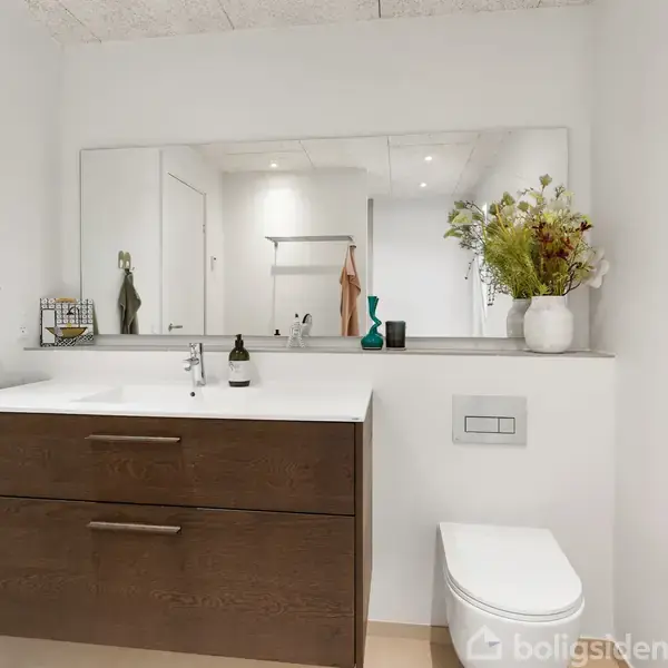 Et badeværelse med hvid vask oven på en mørk skuffeenhed. Vasen med blomster står ved siden af håndvasken. Et spejl fylder væggen over vasken, og der er en toiletpapirholder til højre.
