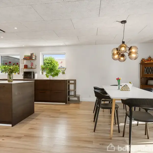 Køkken-alrum med moderne køkkenø i mørkt træ, blomsterdekorationer, spisebord med sorte stole, og en lysende lampe. Lyse trægulve og hvide vægge med vinduer skaber et rummeligt miljø.