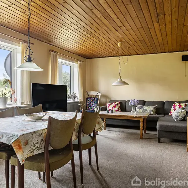 Stue med et spisebord dækket af en blomstret dug ved siden af en grå sofa med farverige puder. To vinduer med gardiner lader naturligt lys ind i rummet.