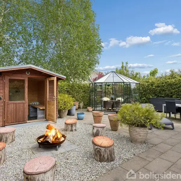 Et bål brænder omringet af træstubbe med puder, foran et træskur i en have med fliseterrasse og drivhus, omgivet af træer og buske under en blå himmel med skyer.