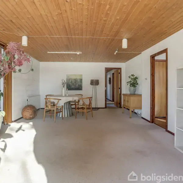 Et lyst rum med et spisebord og stole i midten, omgivet af planter og en hylde til højre. Trægulv og træloft skaber en varm atmosfære.