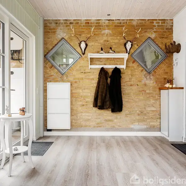 En entre med en rå murstensvæg og lyse trægulve. Jakker hænger på knager over en boks reol. To spejle er diagonalt placeret. Dør og småbord står til venstre.