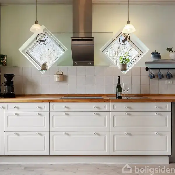 Hvidt køkkenbord med komfur og kaffemaskine, dekorativ flisebaggrund, ophængt bestik, to ruder, hængende lamper. To dekorationskrukker står i vindueskarmen. Flasker og glas står på bordet.