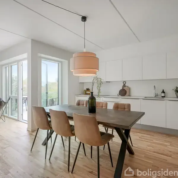 Et spisebord med seks stole står i et moderne køkkenalrum. Der hænger en rund lampe over bordet. Store vinduer til venstre giver udsigt og lys til rummet.