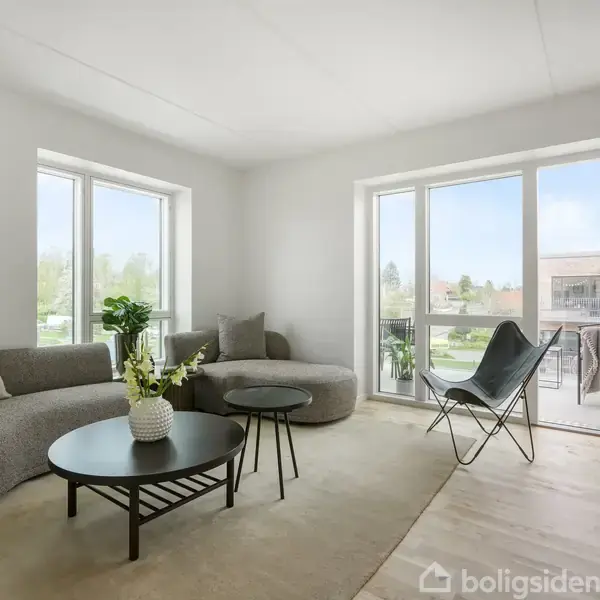 En stue indeholder en grå sofa og et rundt sofabord med blomster. Rummet har trægulv, store vinduer og en åben altandør, der giver udsigt til omgivelserne udenfor.