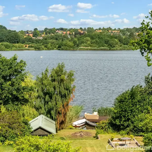 Både ligger stille ved bredden af en sø omgivet af frodig vegetation. I baggrunden ses en bebyggelse på en skovklædt bakke under en blå himmel med spredte skyer.