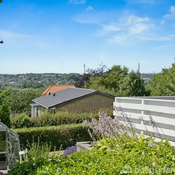En have med blomster og buske står foran et hvidt hegn ved siden af et rødt murstenshus. I baggrunden ses grønne træer, et drivhus og et landskab med bakker.